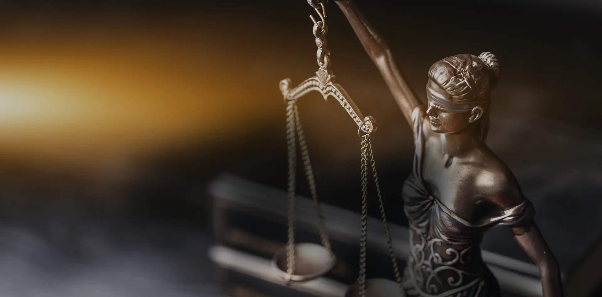 Bronzestatue der Justitia hält eine Waage in der erhobenen rechten Hand, steht in einer schwach beleuchteten Umgebung mit warmem goldenen Licht im Hintergrund. Die Szene vermittelt eine ernste und würdevolle Stimmung und symbolisiert Gerechtigkeit sowie den juristischen Beruf. Kein sichtbarer Text im Bild.